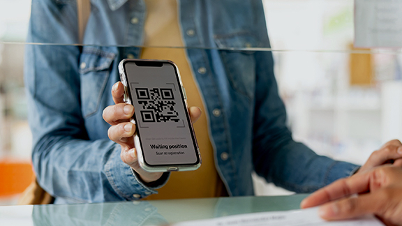 Person zeigt an einem Schalter ein Smartphone mit QR‑Code und digitaler Wartemarken-Anzeige, Symbolbild für die Digitalisierung im öffentlichen Sektor.