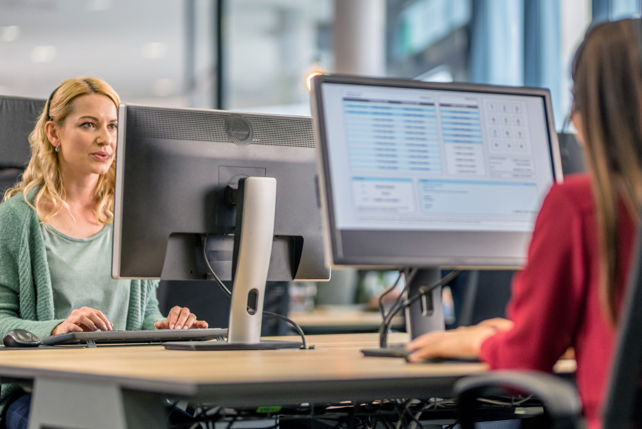 Ein Arbeitsplatz in der öffentlichen Verwaltung Zwei Personen arbeiten an Computerarbeitsplätzen in einem Büro. Im Vordergrund sind Monitore mit geöffneten Anwendungen zu sehen, während eine Person auf einen Bildschirm blickt.