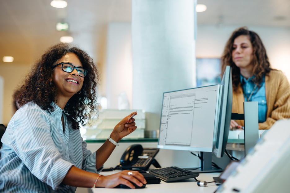 Digitalisierung in der öffentlichen Verwaltung Mitarbeiterin arbeitet an einem Computerarbeitsplatz in einer modernen Büroumgebung, während eine weitere Person im Hintergrund an einem Verwaltungsbereich steht.