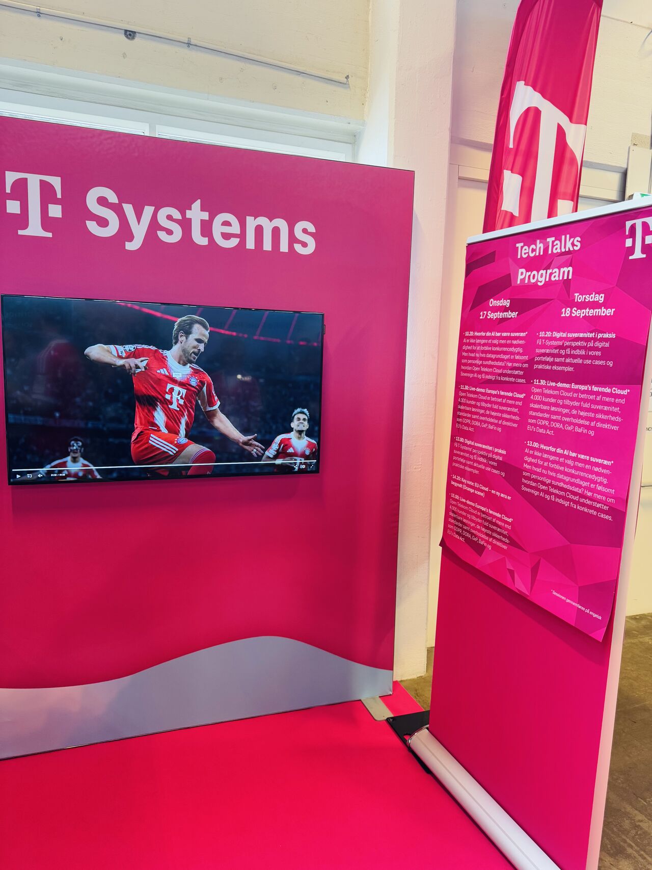 Der T Cloud Public Stand bei mit einem Bildschirm, der ein Fußballspiel zeigt, und einem Veranstaltungshinweis zu den Tech Talks am 17. und 18. September. Der Stand ist in markantem Magenta gehalten.