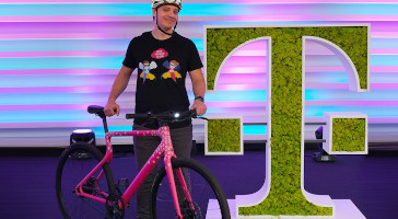 A man is holding a bicycle next to a large Telekom logo covered in moss.