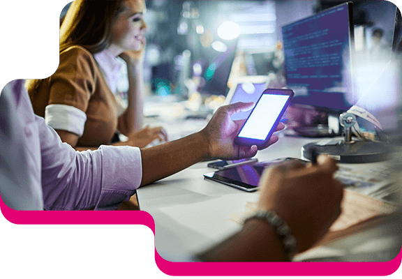 Zwei Personen im Büro arbeiten an Laptop und mit dem Handy, ein Tablet liegt auf dem Tisch und ein Bildschirm steht dahinter.